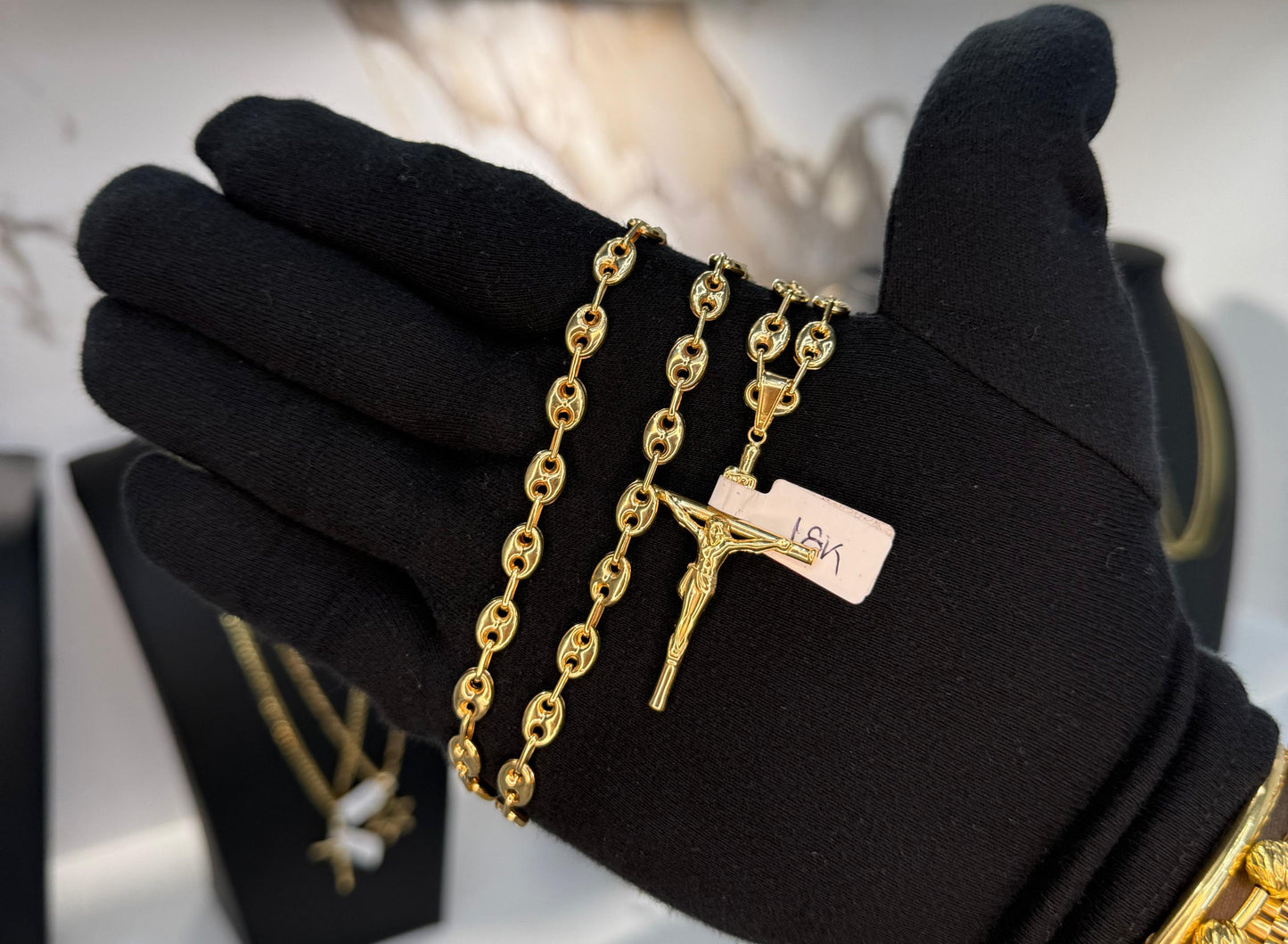 Black glove with gold chain and pendant held by a gloved hand, with jewelry display in the background.